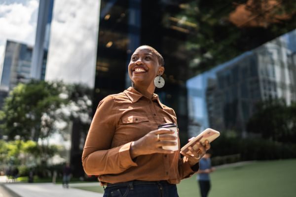 Mulher sorridente segurando um celular e uma bebida, com um fundo urbano, transmitindo confiança e alegria no ambiente de trabalho.