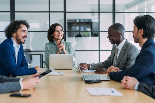 Um grupo diversificado de profissionais em uma reunião, trabalhando juntos e discutindo ideias em uma mesa de conferência moderna, com laptops e dispositivos tecnológicos ao redor.