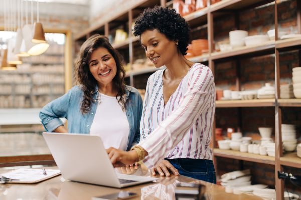 Empresárias sorridentes analisando informações em um laptop dentro de uma loja, representando colaboração e empreendedorismo.