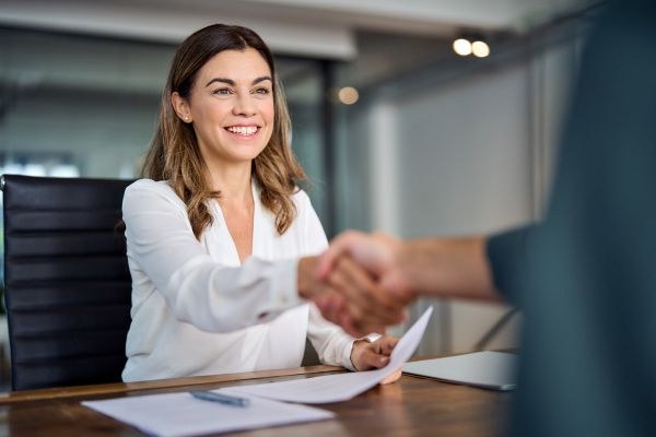 Mulher sorridente em ambiente de escritório, vestindo camisa branca, apertando a mão de outra pessoa, simbolizando uma entrevista ou reunião profissional.