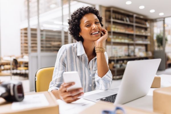 Mulher sorridente de camisa listrada, segurando um celular e sentada em frente a um laptop, em ambiente de trabalho com caixas ao redor, indicando empreendedorismo.