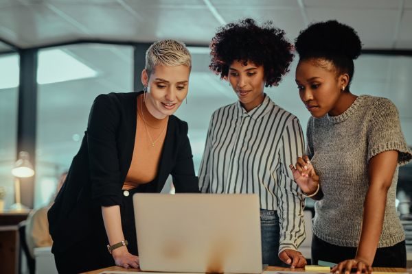 Três mulheres em um ambiente de escritório, colaborando e sorrindo enquanto observam um laptop, demonstrando um momento de troca de ideias.