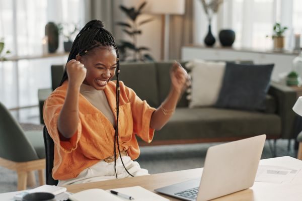 Mulher sorrindo e comemorando com as mãos levantadas enquanto trabalha em um laptop em um ambiente de escritório em casa.