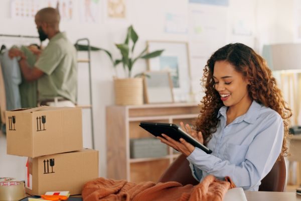 Mulher feliz usando tablet em um ambiente de trabalho criativo, rodeada por caixas e roupas, enquanto homem organiza peças de vestuário ao fundo.